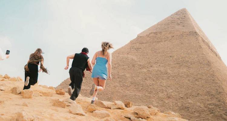 Three travellers run up a sandy slope toward a massive pyramid on a bright day, one person filming with a phone.