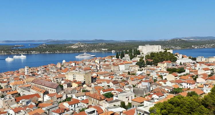 Vue panoramique depuis une colline surplombant la vieille ville de Šibenik, la forteresse et les navires de croisière mouillés dans une baie d'un bleu éclatant.