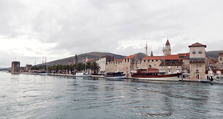 Bord de mer de Trogir avec des bâtiments en pierre, des clochers et des bateaux amarrés sous un ciel couvert.