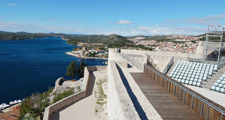Vue depuis une forteresse au sommet d'une colline surplombant l'embouchure d'une rivière, avec des terrasses en pierre et des places assises et une mer bleu vif en contrebas.