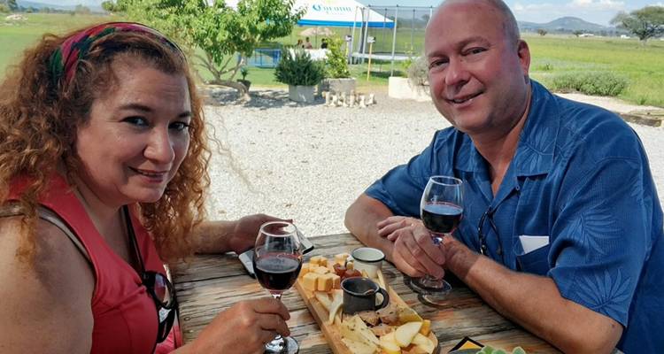 Couple enjoying red wine and a cheese platter at an outdoor wooden table with rural scenery.