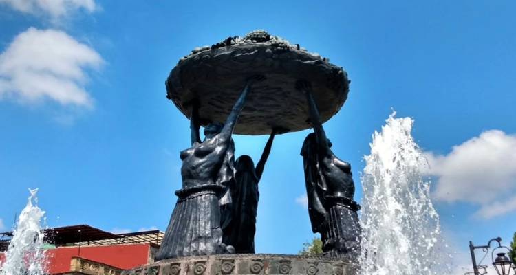 Bronze fountain of three women lifting a large dish with water jets against a bright blue sky in Morelia.