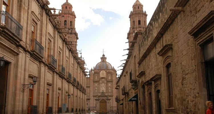 Narrow colonial street lined with stone mansions leading to the domes and twin towers of Morelia Cathedral.
