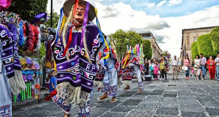 Masked dancers in vibrant woven costumes perform in a lively street parade while crowds watch.