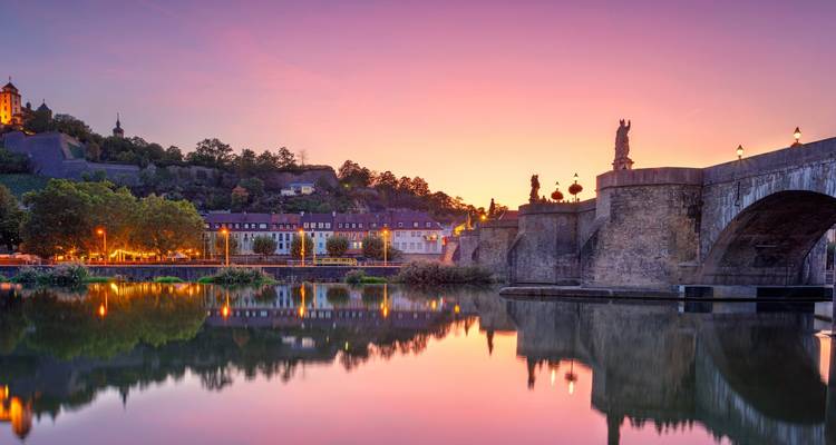 Alte Mainbrücke in Würzburg mit Statuen, die sich gegen einen lebendigen Sonnenuntergangshimmel abzeichnen und im ruhigen Fluss spiegeln.