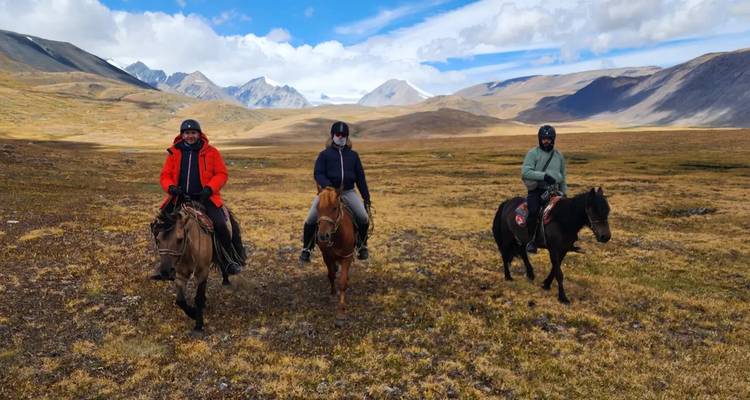 Drei Reiter auf robusten mongolischen Pferden durchqueren eine offene alpine Steppe unter zerklüfteten Gipfeln.