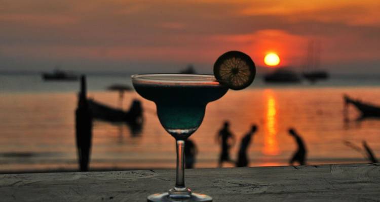 Verre à cocktail coloré au premier plan avec un coucher de soleil orange dramatique et des silhouettes de plage en arrière-plan