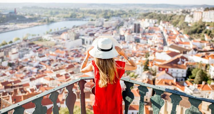 Une femme en robe rouge et chapeau profite d'une vue panoramique depuis un balcon sur les toits de tuiles de Coimbra et la rivière.