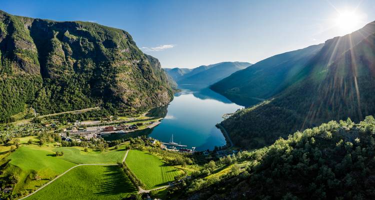 Panorama ensoleillé de l'Aurlandsfjord près de Flåm avec des champs émeraude, des montagnes abruptes et une eau reflétée.