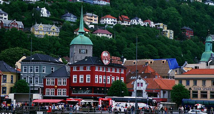 Farbenfrohe Häuser und ein Kirchturm erheben sich vor einem grünen Hang über dem Hafen von Bergen.