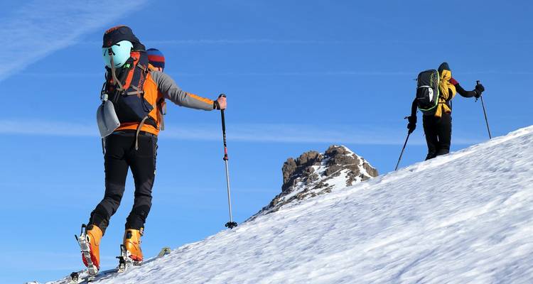 Bergsteiger in heller Ausrüstung steigen einen verschneiten Hang unter einem leuchtend blauen Himmel hinauf.