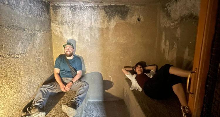 Dos viajeros descansando en antiguos bancos de piedra dentro de una cámara excavada en roca débilmente iluminada.