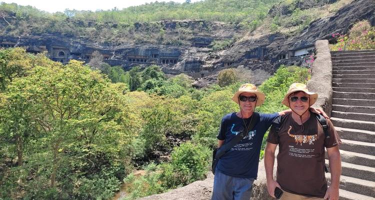 Dos viajeros con sombreros para el sol posan en una vista de escalinata que da a la cara del acantilado boscoso que alberga las entradas de las cuevas de Ajanta.
