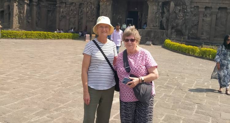 Dos viajeros mayores posando en un gran patio pavimentado ante la ornamentada fachada de un templo excavado en la roca.