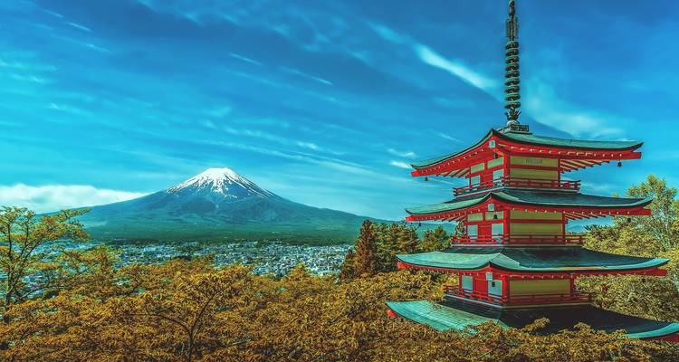 Lebendige rote Chureito-Pagode überblickt den Berg Fuji, der über einer Stadt und Herbstlaub aufragt.