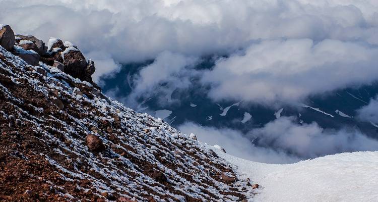 Nahaufnahme eines schneebepuderten Vulkanhangs, der in dichte Wolkendecke abfällt.