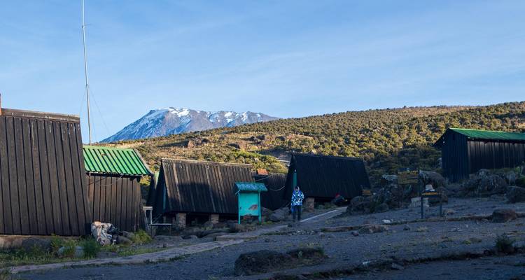 Holzhütten in den Bergen bei Sonnenaufgang mit schneebedecktem Gipfel im Hintergrund und einem einsamen Wanderer in der Nähe.