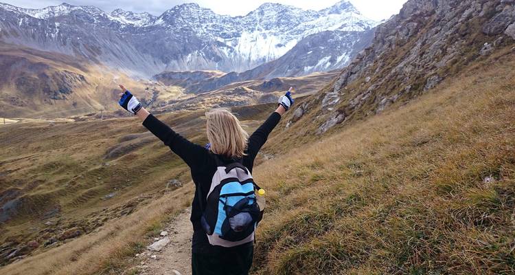 Excursionista mujer levantando los brazos triunfalmente en un sendero de montaña con picos nevados al frente.