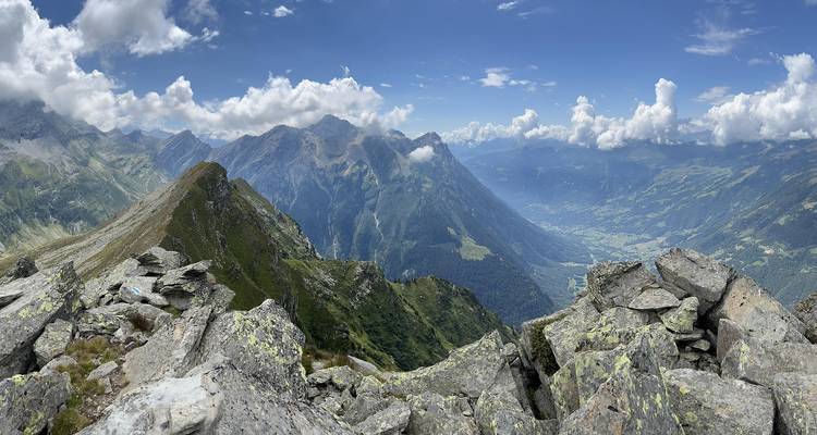 Vista panorámica desde la cumbre rocosa sobre cordilleras alpinas y un valle verde alfombrado de nubes.