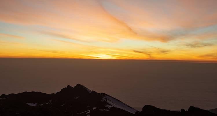 Farbenfroher Sonnenaufgangshimmel über einer dunklen Bergkette und einem Wolkenmeer.