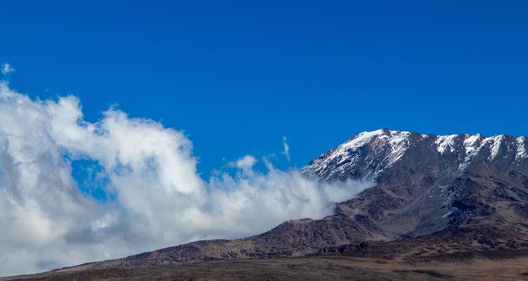 Teilansicht der vulkanischen Hänge des Kilimandscharo mit frischem Schnee vor tiefblauem Himmel und ziehenden Wolken.