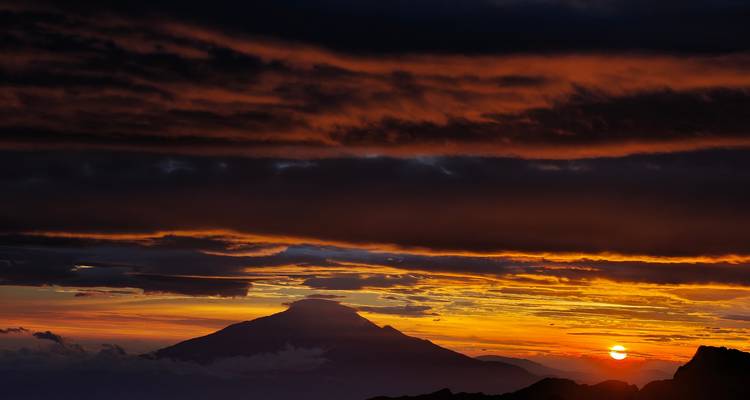 Feurige Sonnenuntergangswolken, die einen kegelförmigen Berggipfel silhouettieren, während die Sonne zum Horizont sinkt.