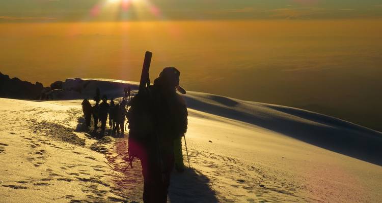 Bergsteiger wandern bei Morgendämmerung über ein leuchtendes Schneefeld unter goldenem Himmel.