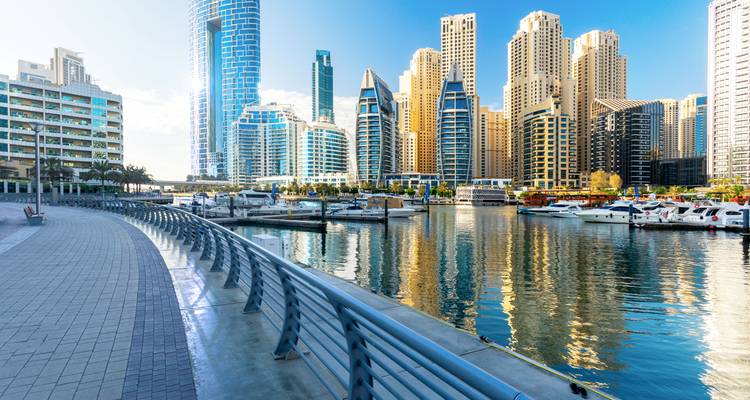 Waterfront von Dubai Marina mit Yachten, Hochhäusern und Spiegelungen in ruhigem Wasser an einem sonnigen Tag.