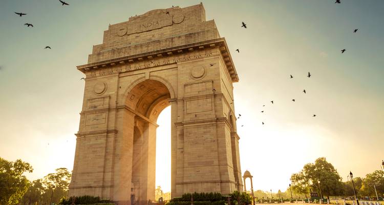 Denkmal India Gate in Silhouette vor einem sanften Sonnenuntergangshimmel mit fliegenden Vögeln.