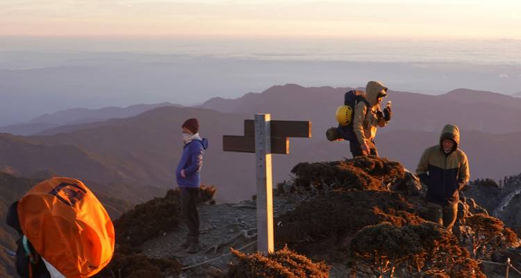 Pequeño grupo de escaladores parados en una cresta rocosa de la cumbre al amanecer con montañas y nubes abajo.