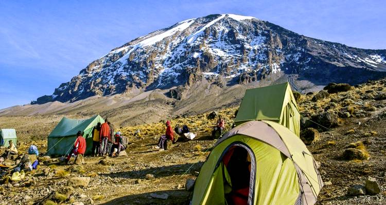 Bunte Zelte und Wanderer lagern auf einem kahlen Hang mit dem schneebedeckten Kilimandscharo, der dahinter unter einem klaren blauen Himmel aufragt