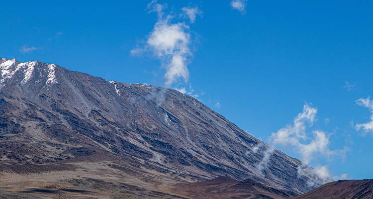 Der felsige, schneebedeckte Hang des Mount Kilimandscharo erhebt sich gegen einen strahlend blauen Himmel mit treibenden Wolken.