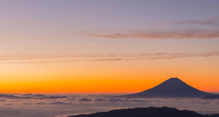 Silhouette eines symmetrischen Vulkangipfels über einem Wolkenmeer unter einem orange-violetten Sonnenaufgangshorizont.