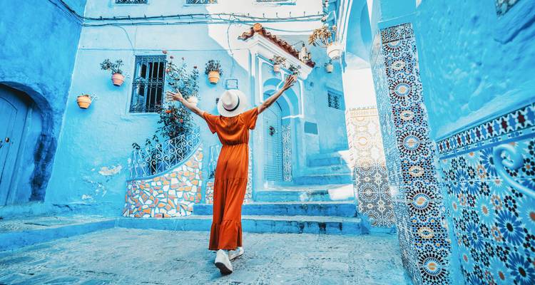 Una mujer con un vestido naranja levanta los brazos alegremente mientras pasea por las calles azules.