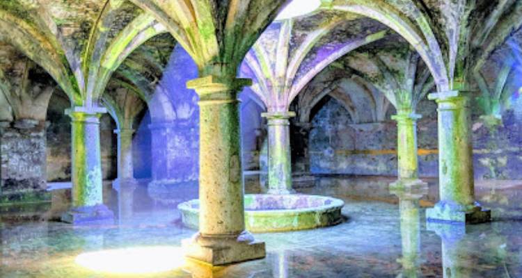 Interior de una cisterna de piedra con columnas y arcos iluminados con luces artificiales de varios colores reflejadas en agua poco profunda.