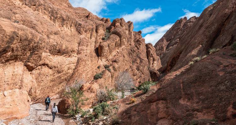 Wanderer gehen durch eine enge rötliche Schlucht mit zerklüfteten Felswänden und blauem Himmel darüber