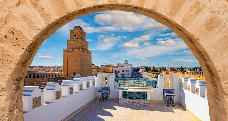 Vue à travers une arche de pierre sur le minaret historique de la Grande Mosquée de Kairouan sous un ciel bleu.