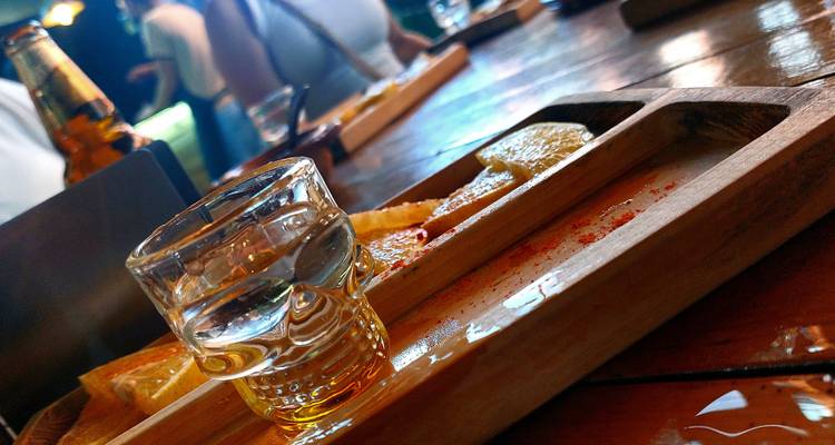 Nahaufnahme eines Mezcal- oder Tequila-Shots auf einem Holztablett mit Salz, Zitrusfrüchten und Freunden im Hintergrund.