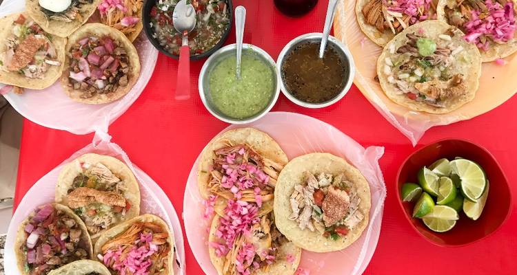 Draufsicht auf verschiedene Tacos mit bunten Toppings, Salsas und Limetten auf einer roten Tischdecke.