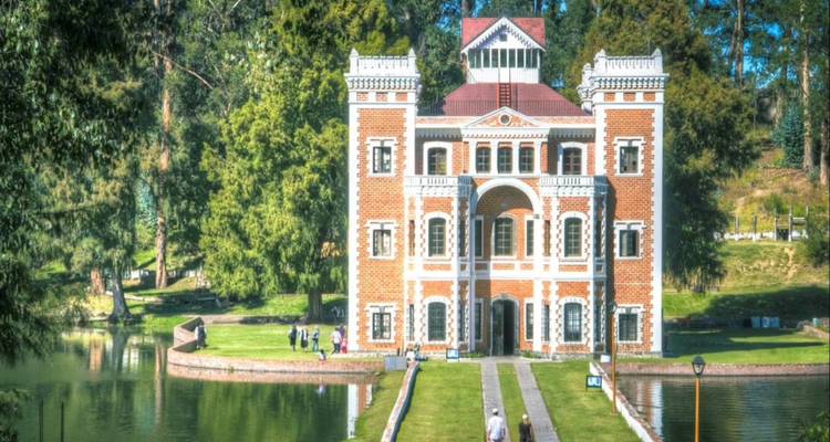 Elegante bakstenen landhuis aan een spiegelende plas omgeven door weelderig bos, bezoekers wandelen langs het pad.