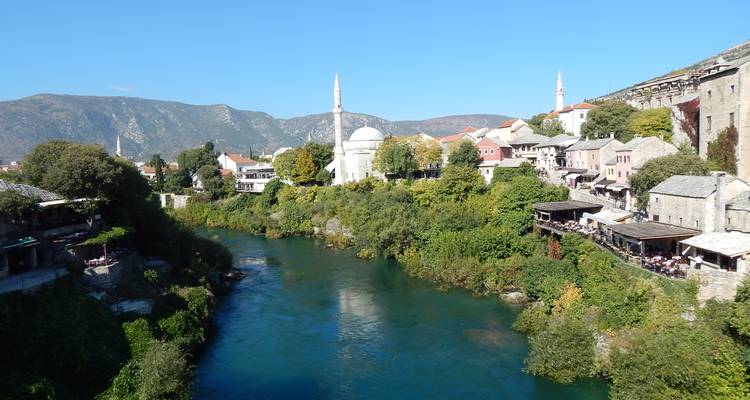 Moskee met witte koepel en slanke minaret met uitzicht op de rivier de Neretva omzoomd met groene bomen.