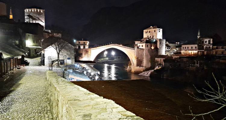 Stari Most brug gloeiend onder nachtverlichting weerkaatst op de Neretva rivier met kasseienpad op de voorgrond.