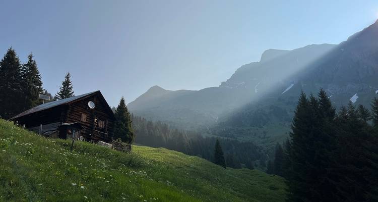 Kleine Holzhütte auf einem grasbewachsenen Hang mit dramatischem Sonnenstrahl, der bei Morgendämmerung durch den Bergdunst schneidet.