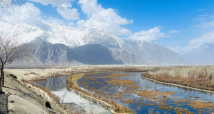 Large rivière serpentant à travers une haute vallée himalayenne avec des sommets enneigés et un ciel bleu.