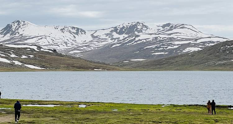 Lac alpin cerné de neige avec des visiteurs au loin se promenant le long de la rive herbeuse sous un ciel couvert.