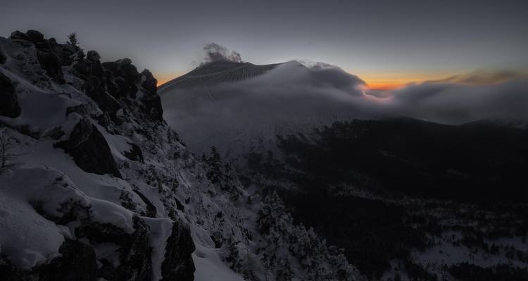 Schnebedeckte Vulkankämme fallen in die Dunkelheit ab, während das Morgenlicht den Horizont hinter einem dampfenden Krater färbt.