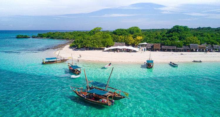 Kristallklares türkisfarbenes Wasser umgibt eine Insel mit weißem Sand, auf der traditionelle hölzerne Dhau-Boote verstreut sind.
