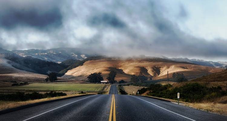 Leere Asphaltstraße erstreckt sich zu sanften goldenen Hügeln, die teilweise in Nebel gehüllt sind