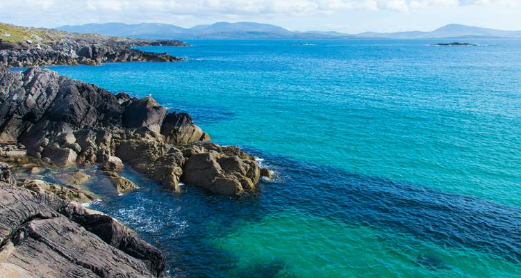 Littoral rocheux avec une eau turquoise cristalline et des montagnes lointaines sous un ciel lumineux.
