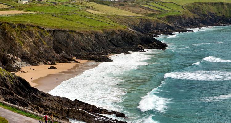 Les vagues se brisent sur une crique sablonneuse isolée sous des champs en terrasses verdoyants et des falaises rocheuses.